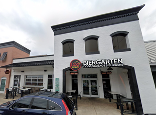 Exterior of a building with a sign reading "Dog Haus Biergarten Dogs, Sausages, Burgers." Outdoor seating and a parked car in front.