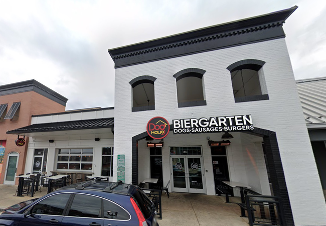 Exterior of a building with a sign reading "Dog Haus Biergarten Dogs, Sausages, Burgers." Outdoor seating and a parked car in front.