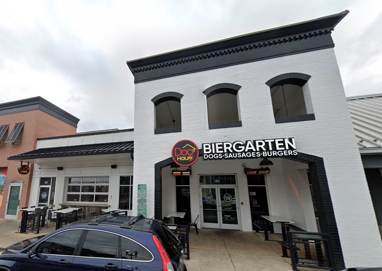 Exterior of a building with a sign reading "Dog Haus Biergarten Dogs, Sausages, Burgers." Outdoor seating and a parked car in front.