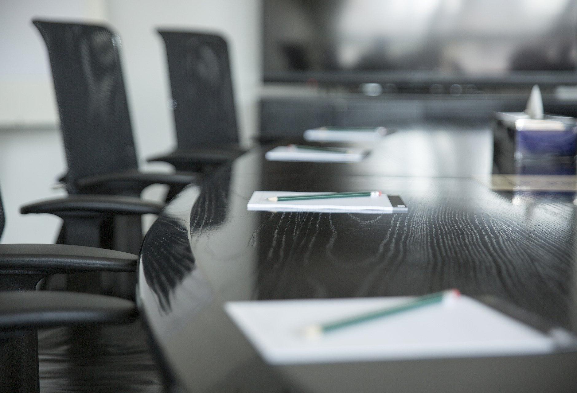 A large boardroom table with empty chairs. There are notebooks and pencils on the table but no people in the frame.