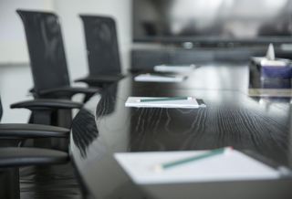 A large boardroom table with empty chairs. There are notebooks and pencils on the table but no people in the frame.