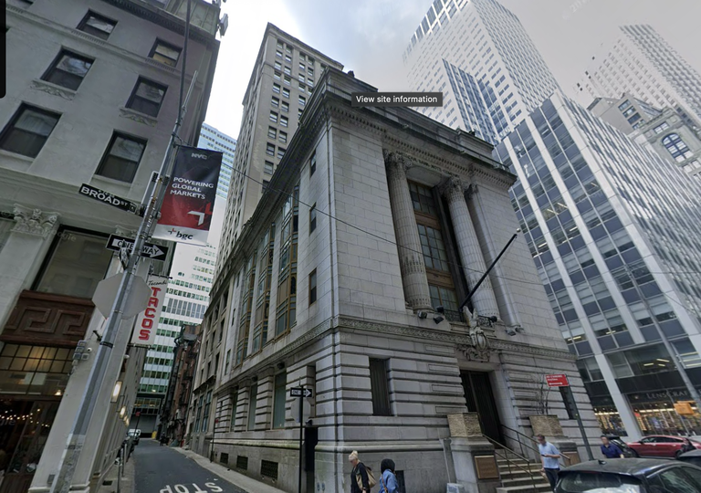 Street view of a historic building with columns on a corner, surrounded by tall modern buildings. People walk on the sidewalk below.