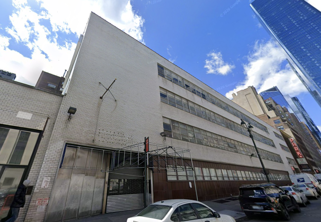 A long, beige brick building with rows of windows, situated in an urban area, under a partly cloudy blue sky, with parked cars along the street.