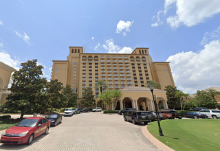 Tall beige hotel with balconies and a large entrance, surrounded by cars and palm trees under a partly cloudy sky.