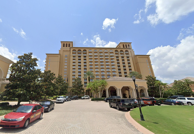 Tall beige hotel with balconies and a large entrance, surrounded by cars and palm trees under a partly cloudy sky.
