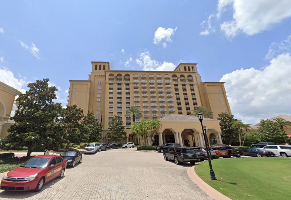 Tall beige hotel with balconies and a large entrance, surrounded by cars and palm trees under a partly cloudy sky.