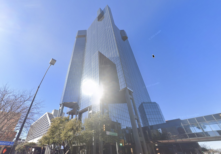 Tall glass skyscraper reflecting sunlight, set against a clear blue sky, with trees and a street sign visible at its base.