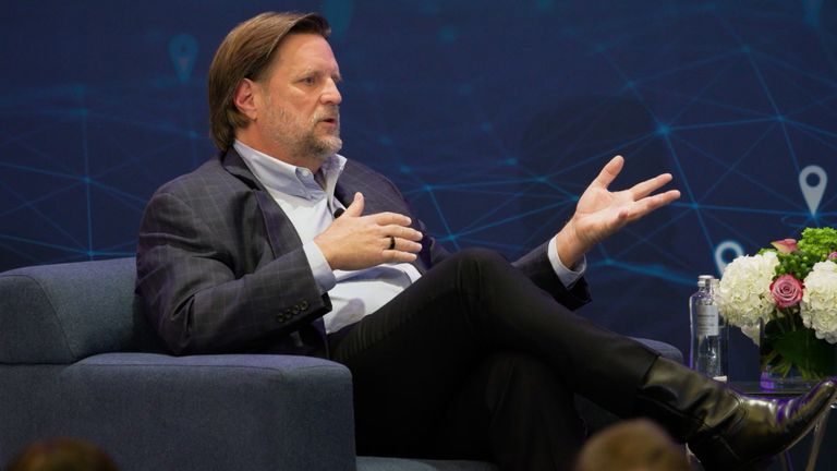 Man in a suit sits on a chair, gesturing during a discussion. Behind him is a blue patterned backdrop. A bouquet and a bottle of water are nearby.
