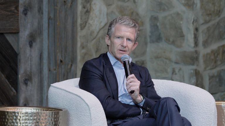 Man in a suit talking into a microphone while seated in a white chair against a stone wall backdrop.