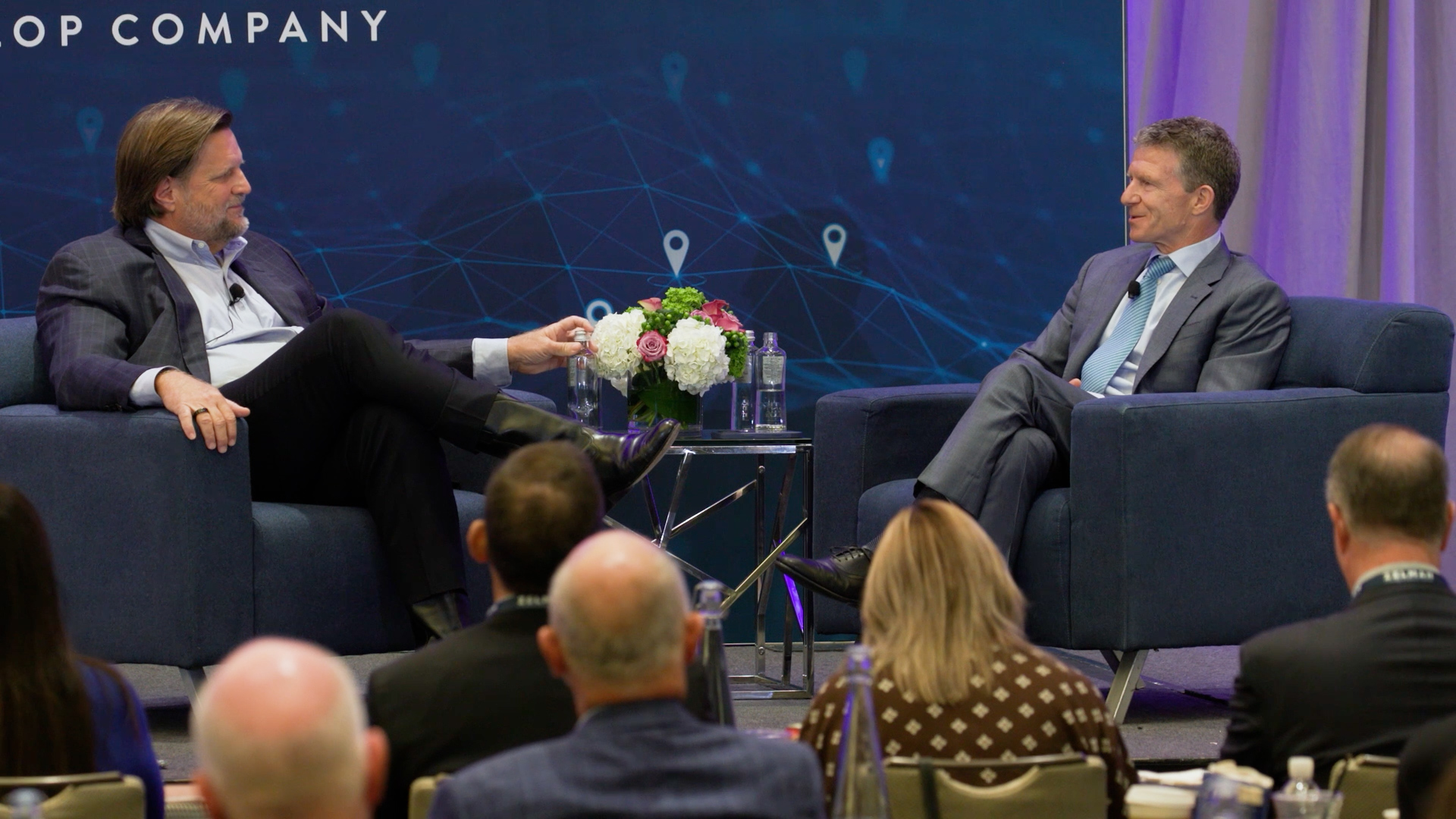 Two men in suits sit on stage in a discussion, with a floral centerpiece between them. An audience is visible in the foreground.