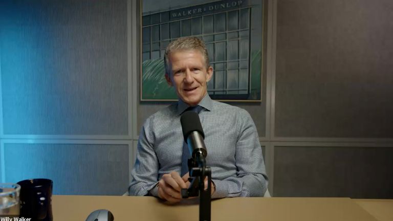 A man in a checkered shirt sits at a table with a microphone in front of him, smiling, with a framed picture in the background.