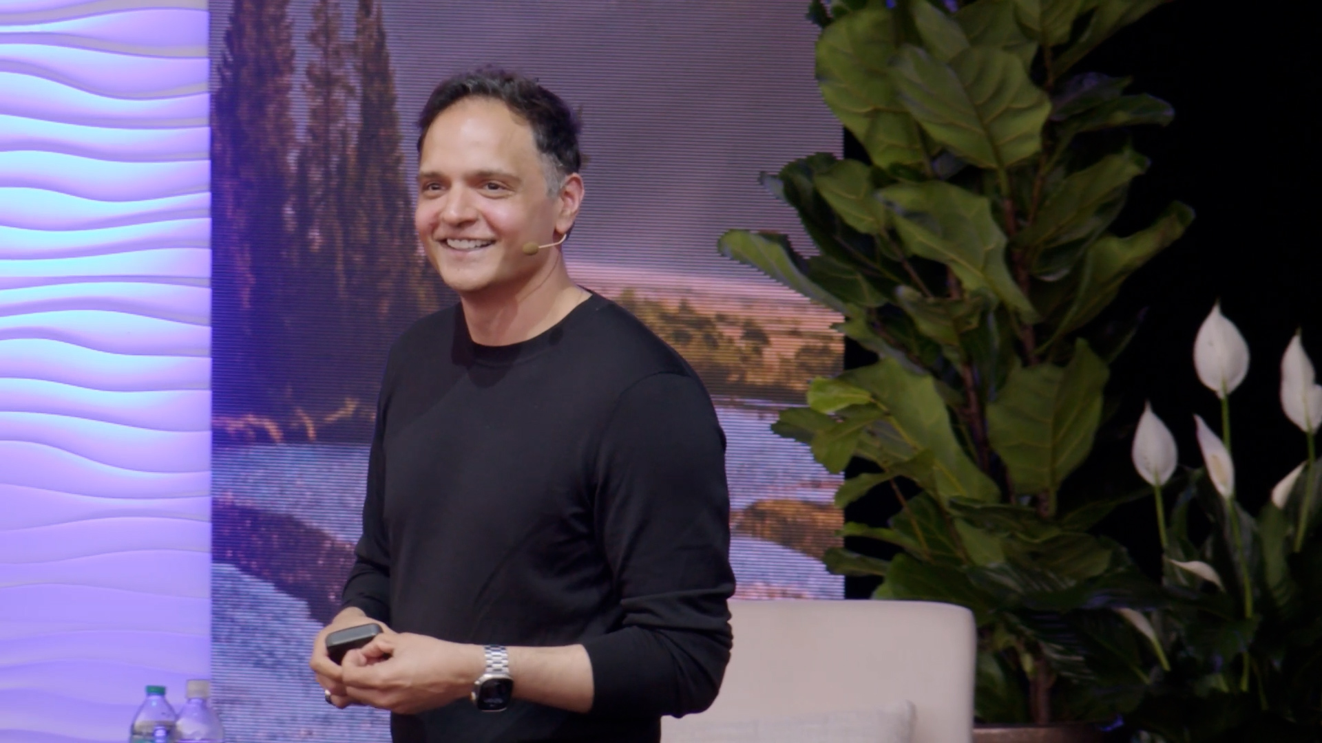 A smiling man in a black shirt speaks on stage with a headset mic, standing near plants and a textured wall backdrop.