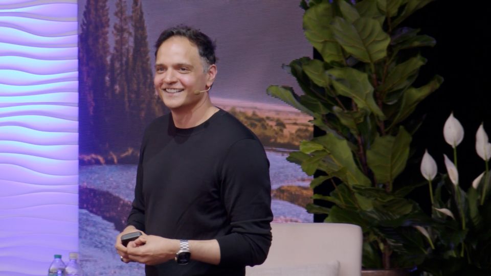 A smiling man in a black shirt speaks on stage with a headset mic, standing near plants and a textured wall backdrop.