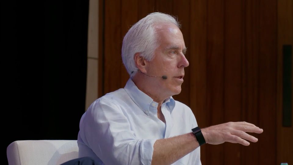 Older man with white hair, wearing a light blue shirt, speaking into a headset microphone and gesturing with his hand.