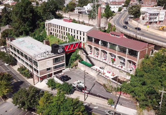 Aerial view of industrial lofts with red bridge, adjacent to a road and surrounded by trees and buildings. Parking lot visible below.