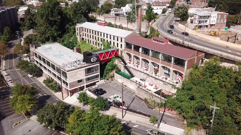 Aerial view of industrial lofts with red bridge, adjacent to a road and surrounded by trees and buildings. Parking lot visible below.