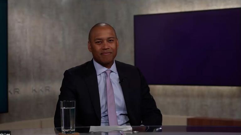 A man in a suit and tie sits at a desk with a glass of water, smiling, with a grey and purple background.