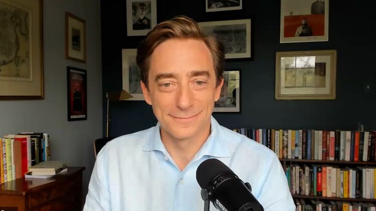 Person in a light blue shirt smiling, sitting in a room with framed pictures and a bookshelf, speaking into a black microphone.