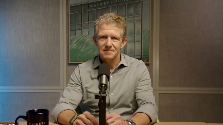 A man sits at a table with a microphone, wearing a grey shirt. A framed picture and a mug are visible in the background.