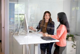 An image of two women in an office enclosed by glass in front of what appears to be a Mac computer.