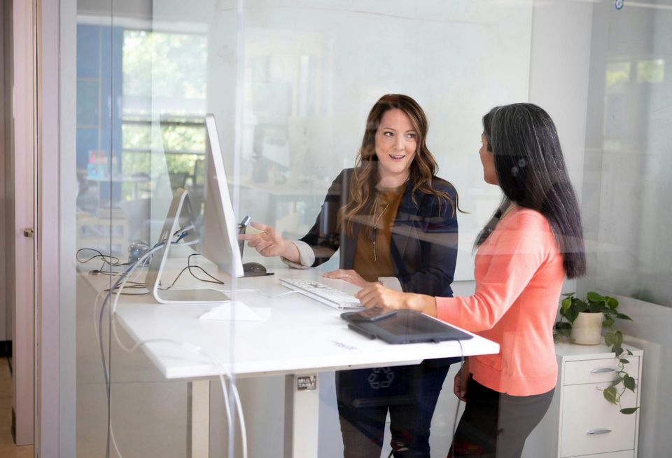 An image of two women in an office enclosed by glass in front of what appears to be a Mac computer.