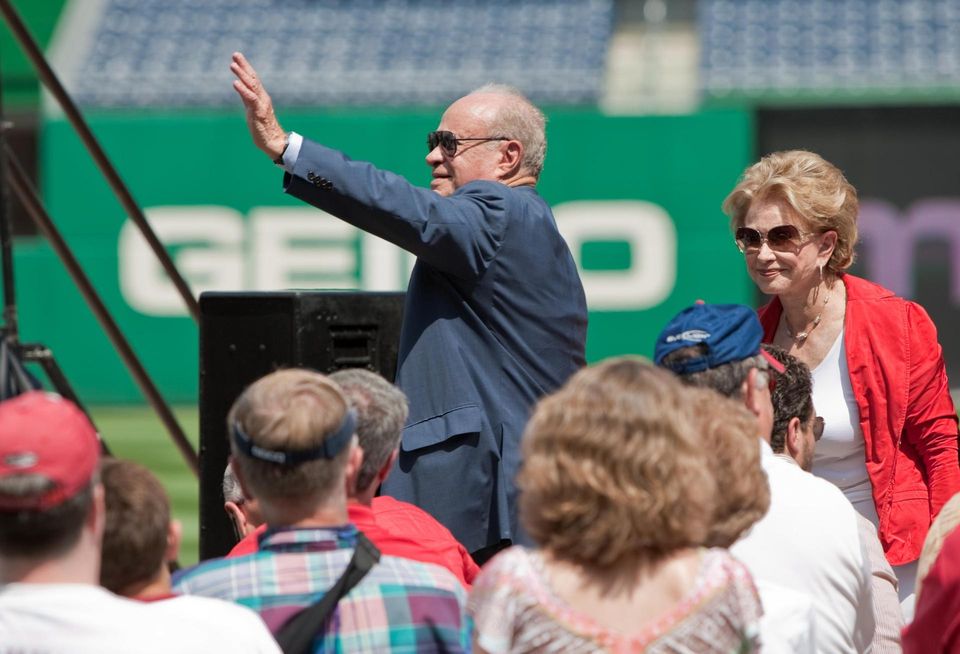 Ted Lerner in suit stands and waves in a baseball stadium with his wife, Annette, standing nearby