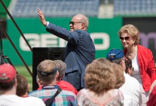 Ted Lerner in suit stands and waves in a baseball stadium with his wife, Annette, standing nearby