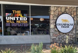 A photo of the storefront at Champagne Tiger, with a sign in the window that reads "The people united will never be defeated" in various colors and fonts.