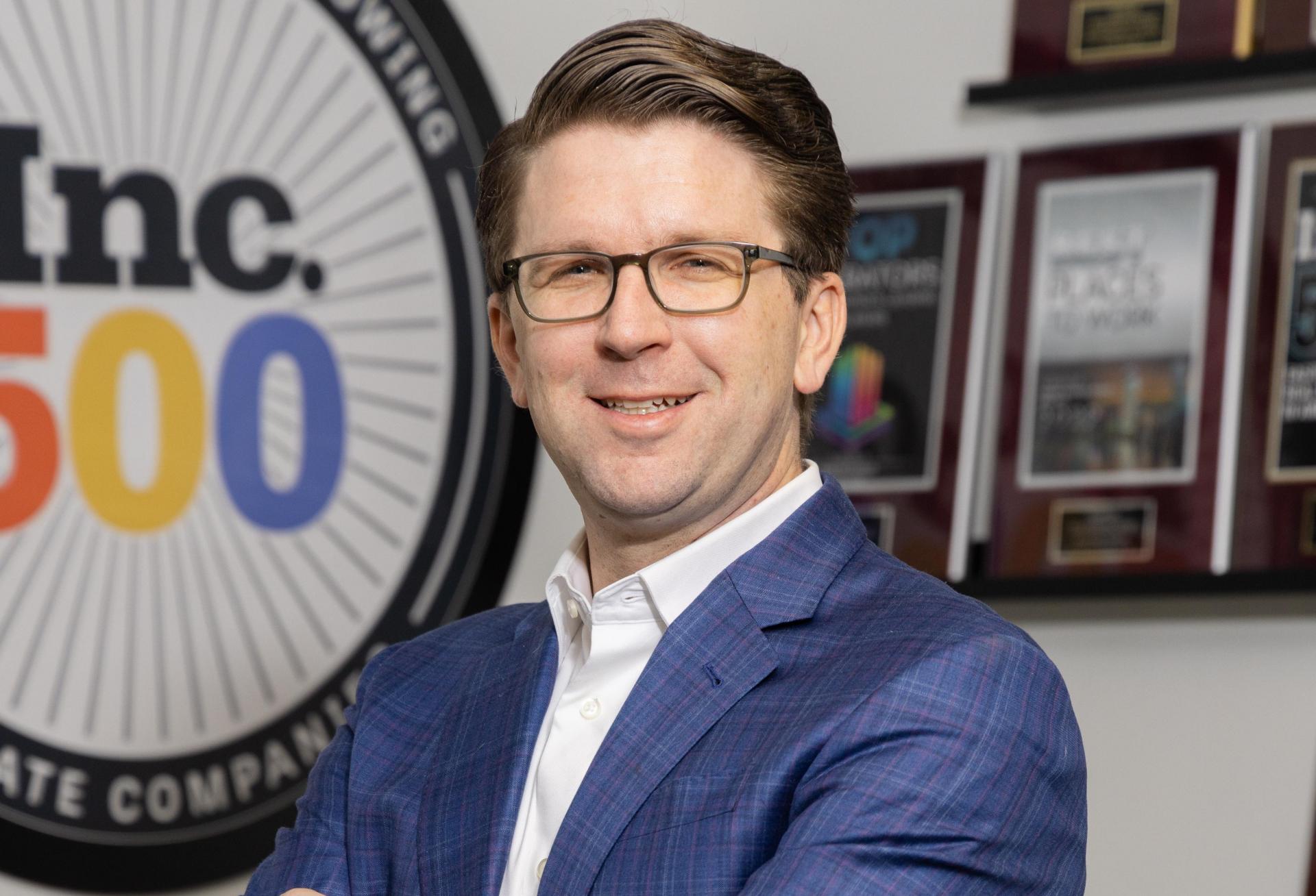 A professional portrait of Spartan Investment Group co-founder, president and chief investment officer Ryan Gibson in a blue suit standing in his office.