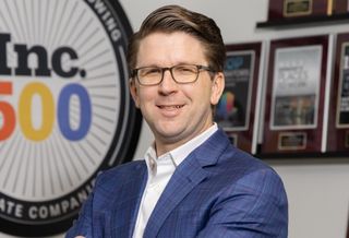A professional portrait of Spartan Investment Group co-founder, president and chief investment officer Ryan Gibson in a blue suit standing in his office.