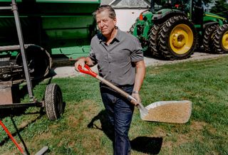 Farmland Partners Founder and Chairman Paul Pittman working on a farm with a shovel, seeds and tractors in the background.