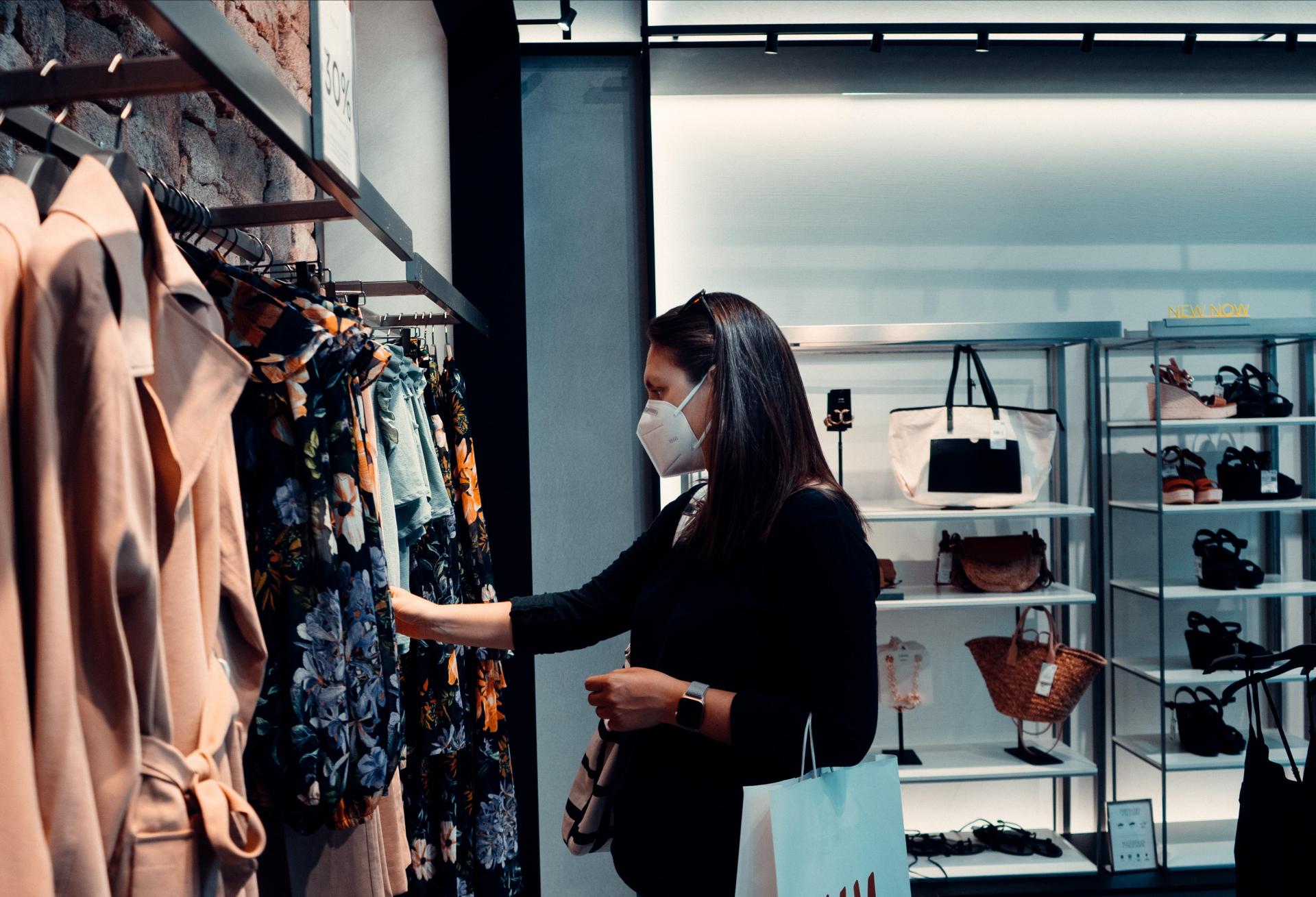 A woman shops for clothes wearing a mask. No other people re visible in the frame. 