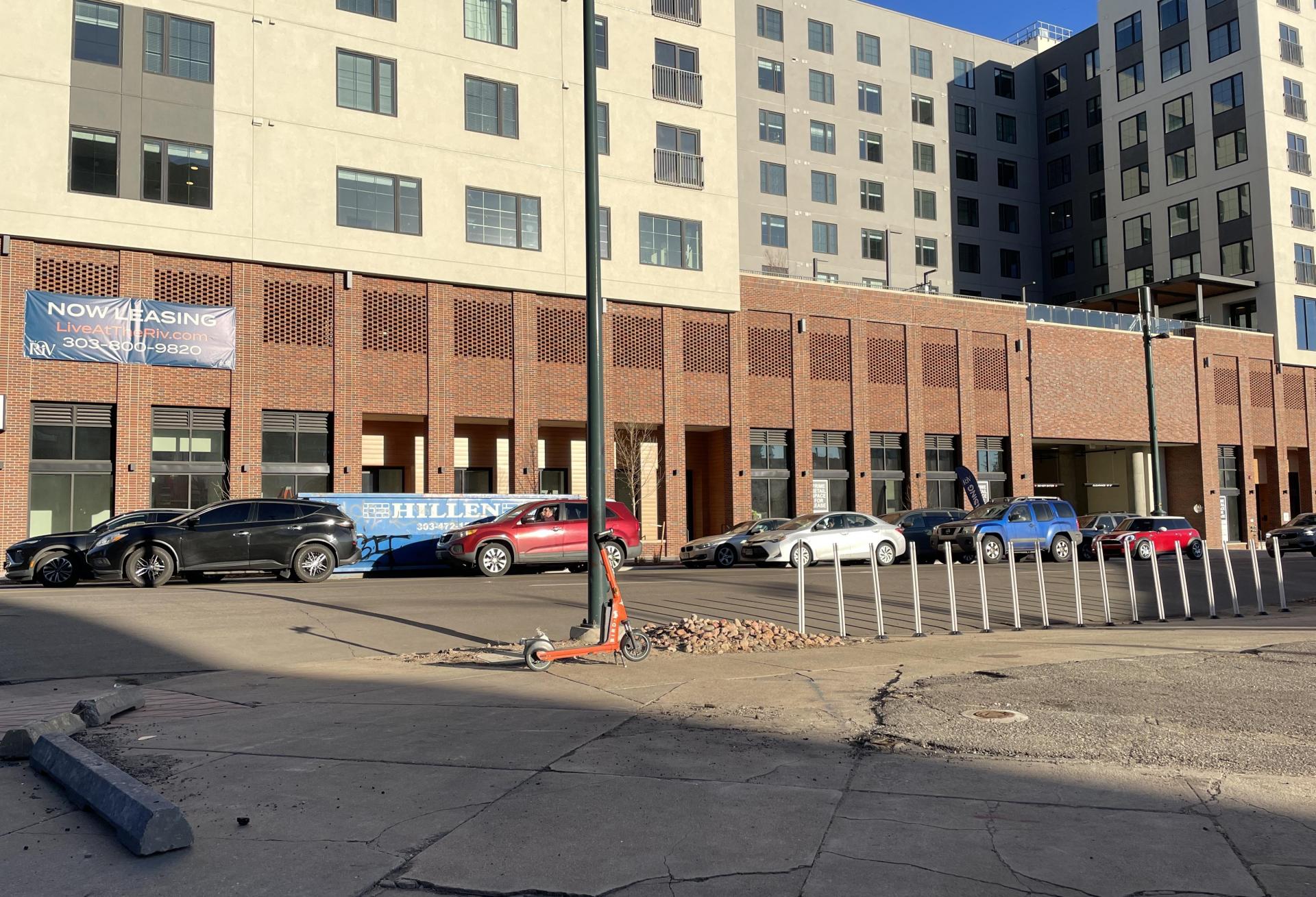 An image of The Riv multifamily community at rush hour from across Downing Street showing crumbling sidewalks and a dumpster in front of the building, as well as a line of cars waiting to get to the interstate.