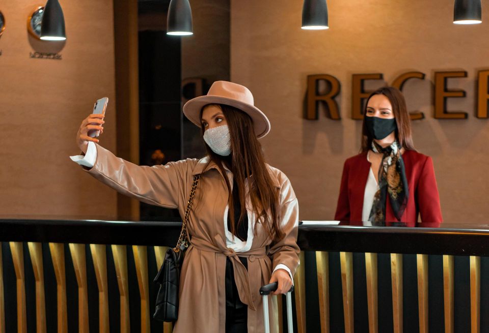 A masked woman takes a selfie in front of a hotel reception desk.