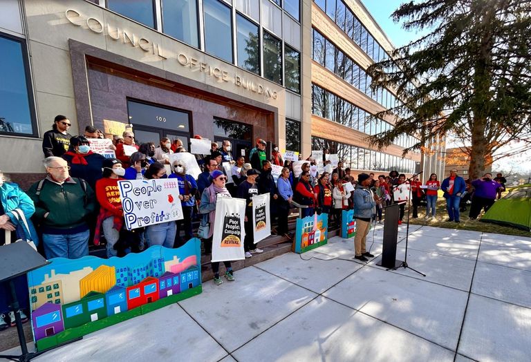 Protesters stand on the steps of a building behind a lectern with speaker. Signs read 