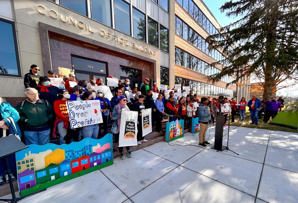 Protesters stand on the steps of a building behind a lectern with speaker. Signs read