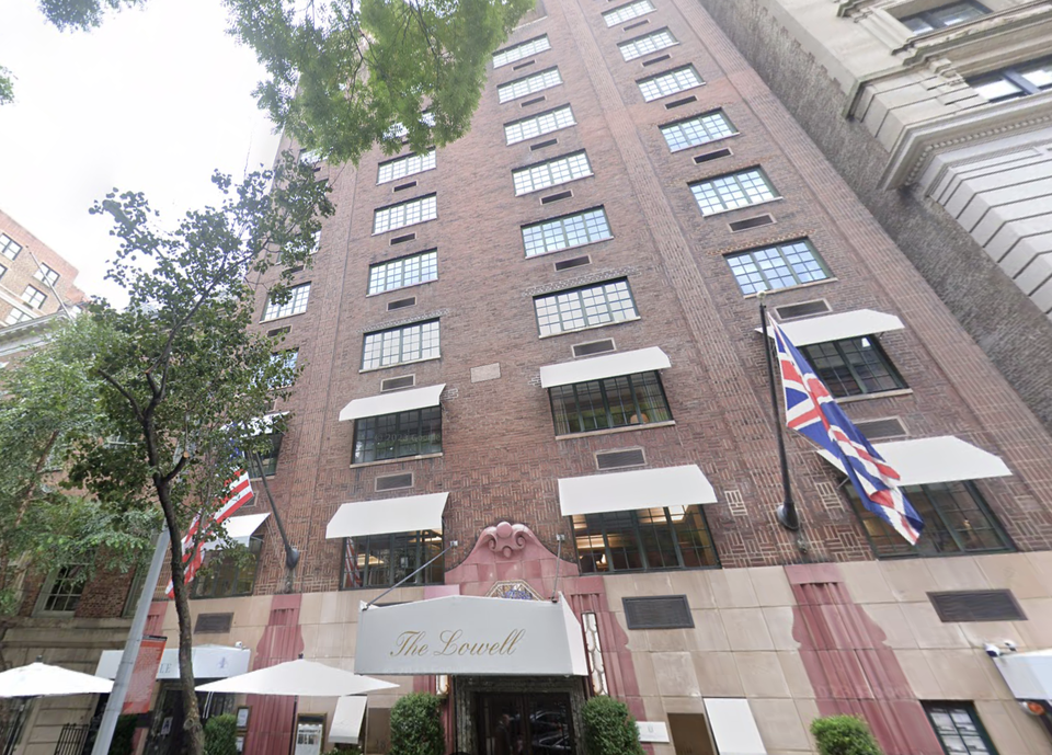 Brick hotel entrance with white awnings, flags, and name "The Lowell" above. Tall building with many windows and trees nearby.