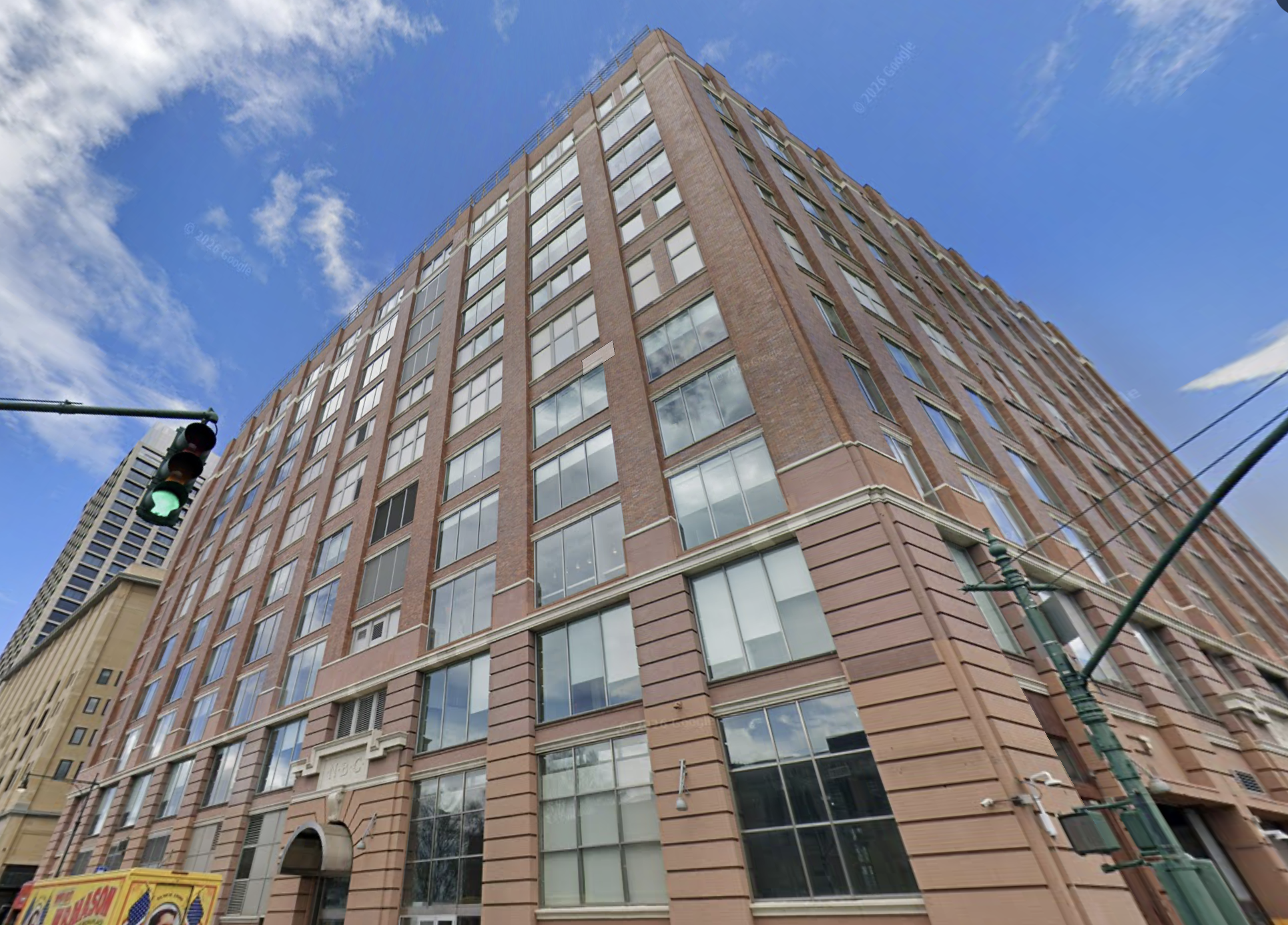 A tall brick building with numerous windows against a blue sky, viewed from a low angle. A green traffic light is visible on the left.