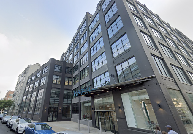 A modern multi-story building with large windows and a black façade, located along a street with parked cars. The entrance has a glass canopy.