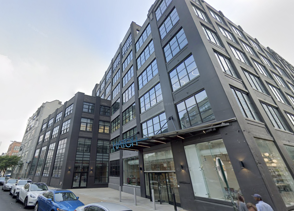 A modern multi-story building with large windows and a black façade, located along a street with parked cars. The entrance has a glass canopy.
