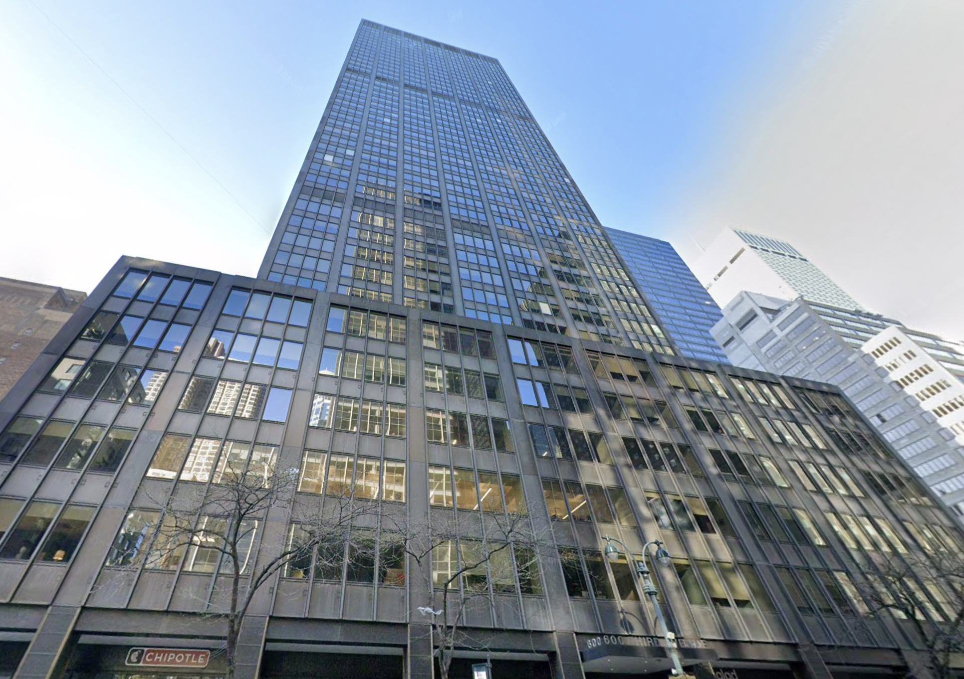 Tall building with a glass façade reflecting the sky and other buildings, viewed from street level. Tree branches and a Chipotle sign are visible below.
