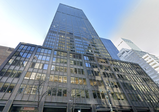 Tall building with a glass façade reflecting the sky and other buildings, viewed from street level. Tree branches and a Chipotle sign are visible below.