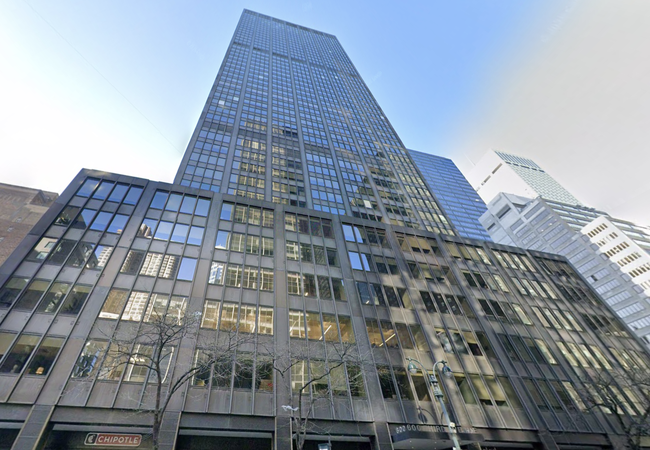 Tall building with a glass façade reflecting the sky and other buildings, viewed from street level. Tree branches and a Chipotle sign are visible below.