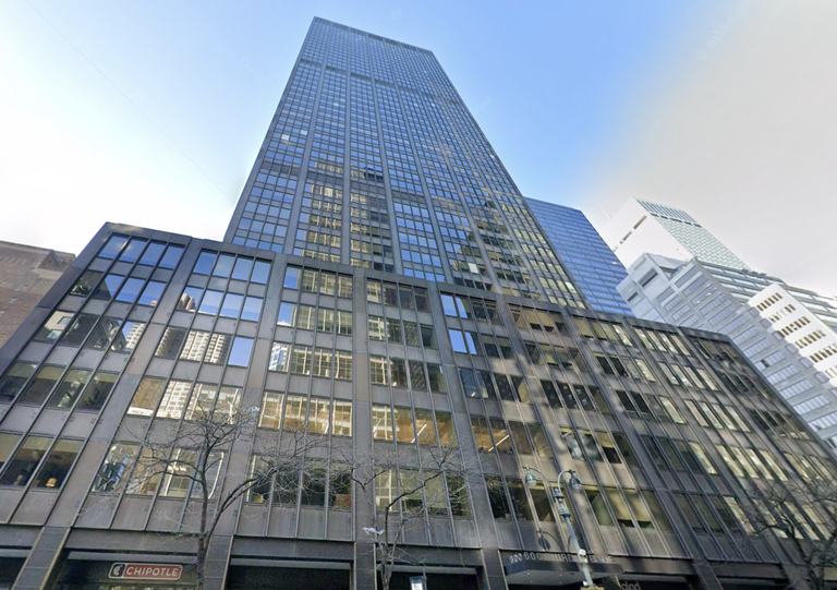 Tall building with a glass façade reflecting the sky and other buildings, viewed from street level. Tree branches and a Chipotle sign are visible below.