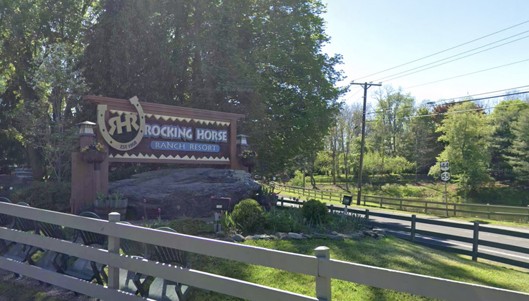 Entrance sign of Rocking Horse Ranch Resort by a road, surrounded by trees and a wooden fence, in a rural setting.
