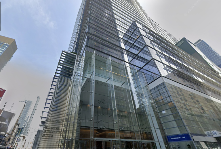 Tall modern skyscraper with a glass facade, reflecting the sky, with Bluestone Lane signage at the ground level. Nearby buildings visible.