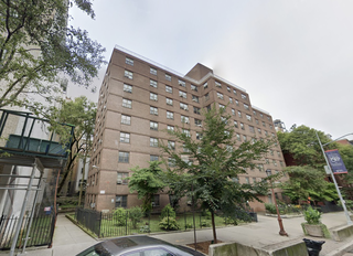Brick apartment building with multiple stories, surrounded by trees and a black metal fence. Sidewalk and a car in the foreground.
