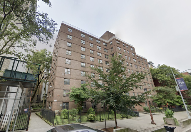 Brick apartment building with multiple stories, surrounded by trees and a black metal fence. Sidewalk and a car in the foreground.