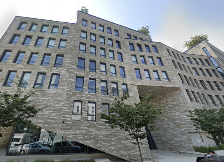 Modern, multi-story brick building with large windows, slanted base, and trees on the sidewalk and rooftop, against a clear sky.