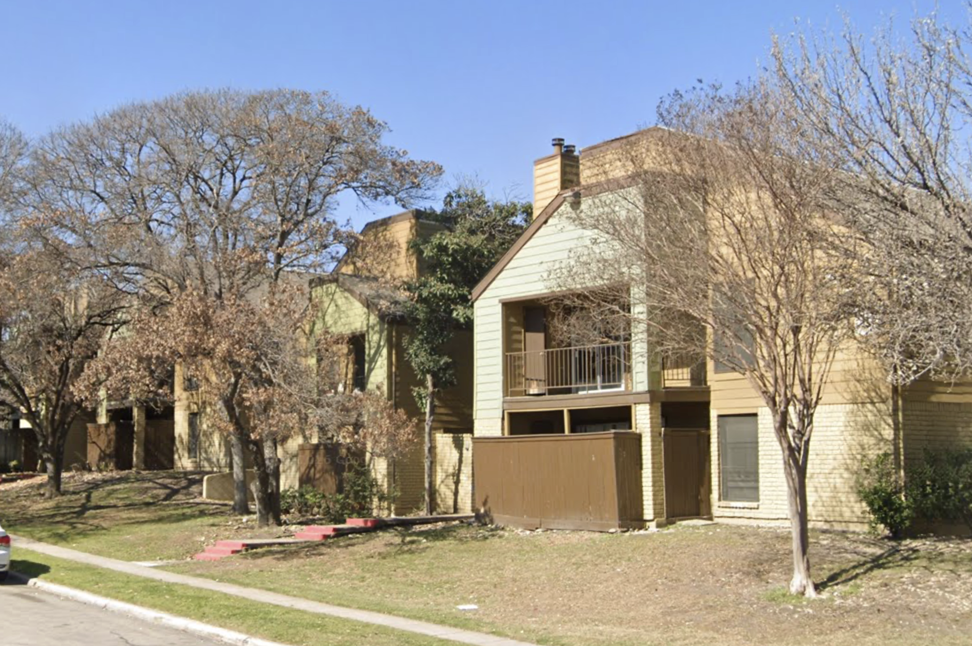 Two-story apartment building with a balcony, surrounded by leafless trees and a grassy area, under a clear blue sky.